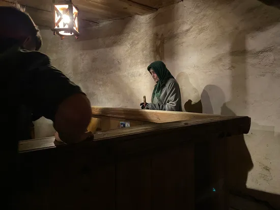 Woman in medieval clothing working with tools under lantern light inside rustic wooden workshop