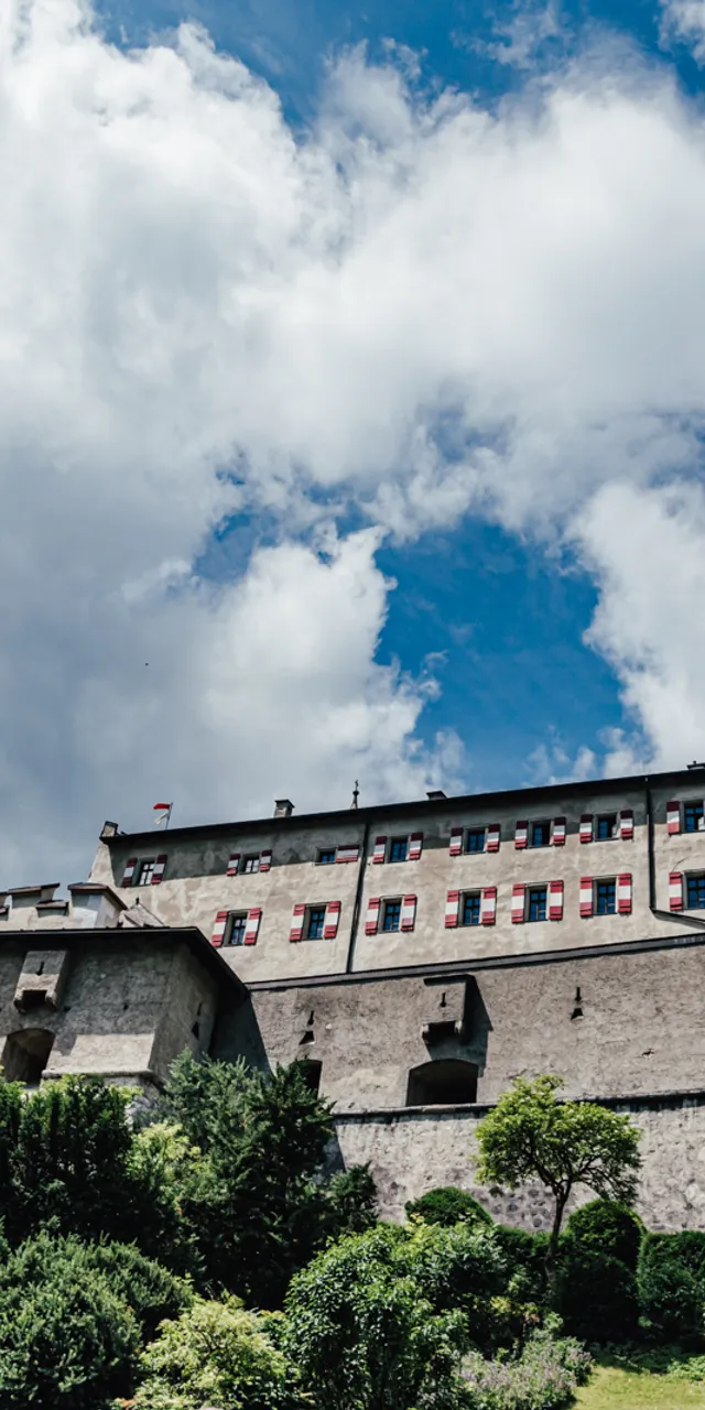 Historic stone castle with red-shuttered windows and towers under a cloudy blue sky