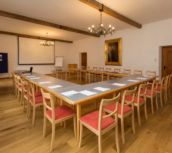 Konferenzraum mit U-förmiger Tischanordnung, roten Stühlen, Projektionsleinwand und Kronleuchtern