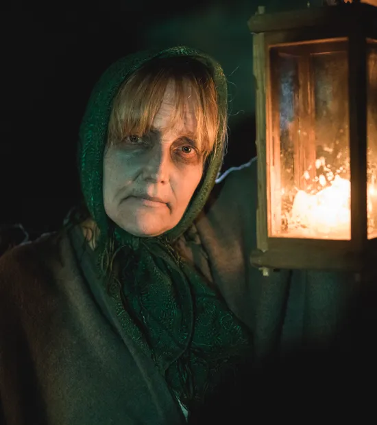 Woman in dark cloak and green headscarf holding lit lantern in low light